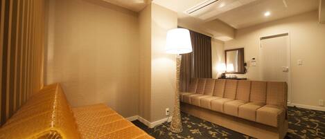 A hotel room with a sofa, a patterned bedspread, a standing lamp, and a carpeted floor.