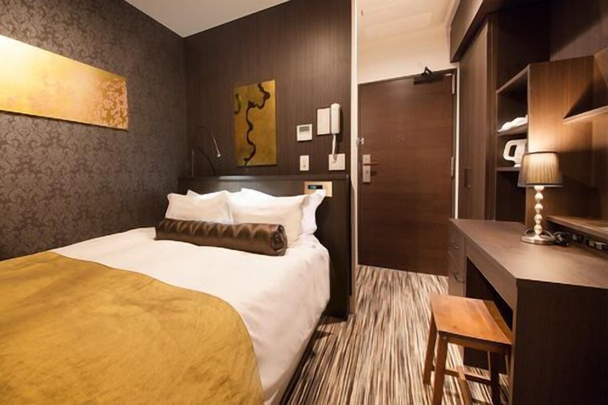 A hotel room with a bed, a desk, a chair, a lamp, and a painting on the wall.