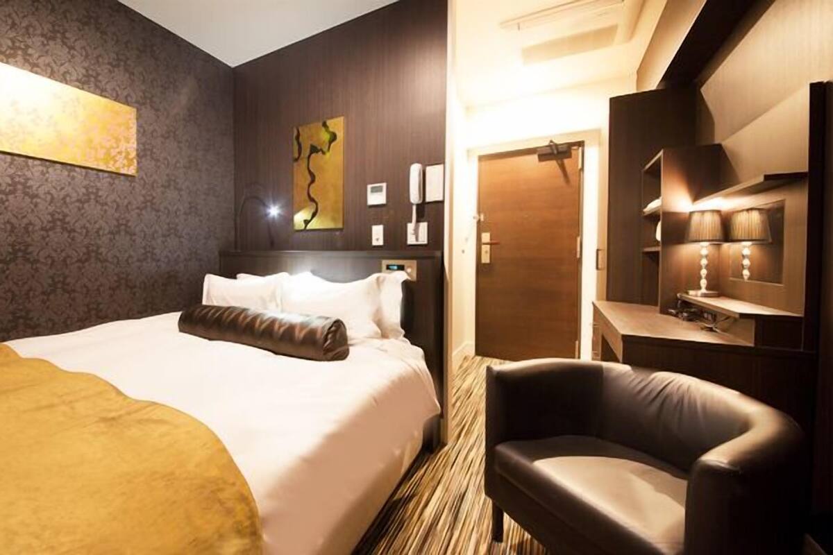 A hotel room with a large bed, a chair, a desk with a lamp, and a door.