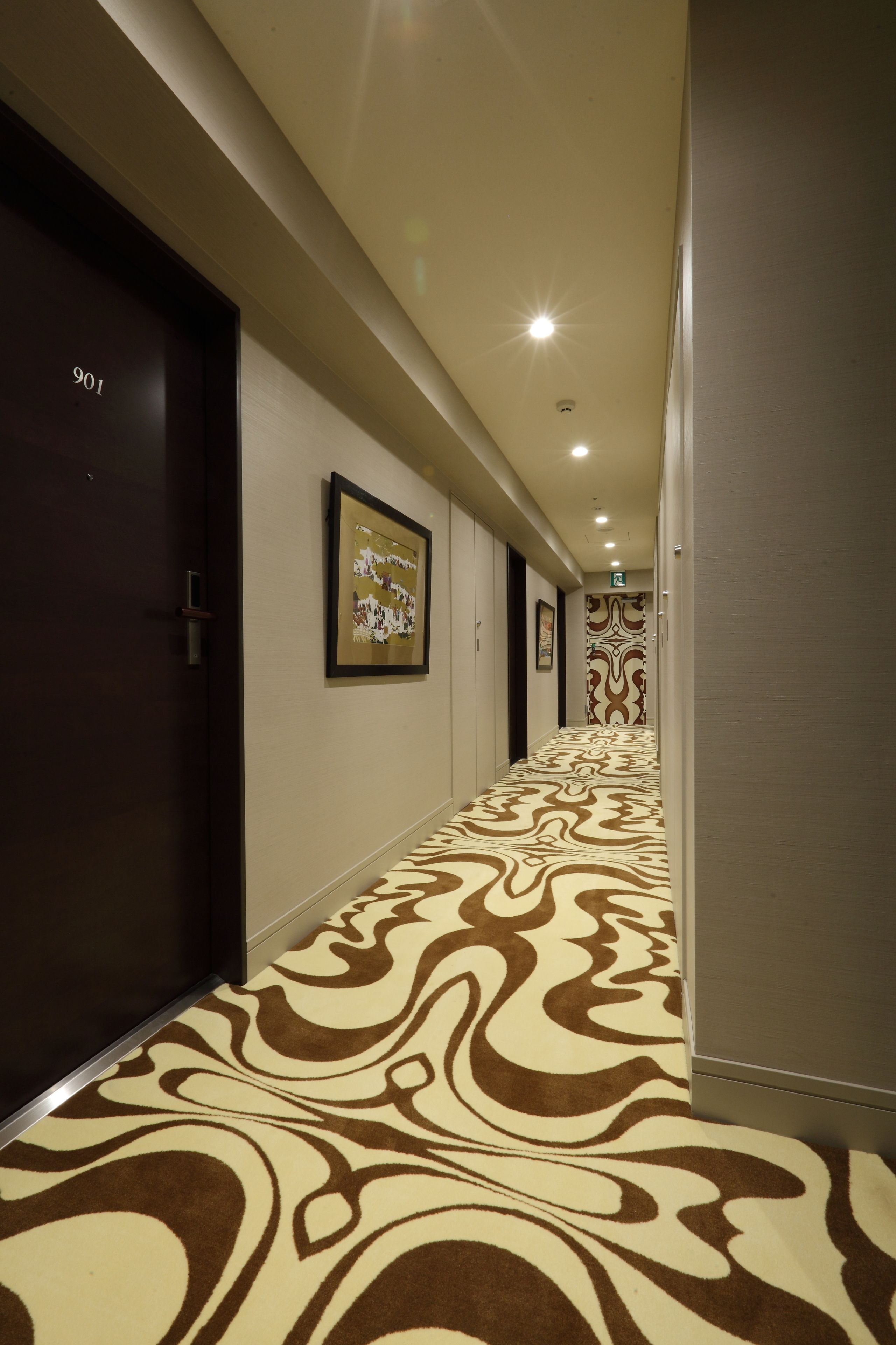 A hotel corridor with patterned carpet, dark doors, and framed artwork on the walls.