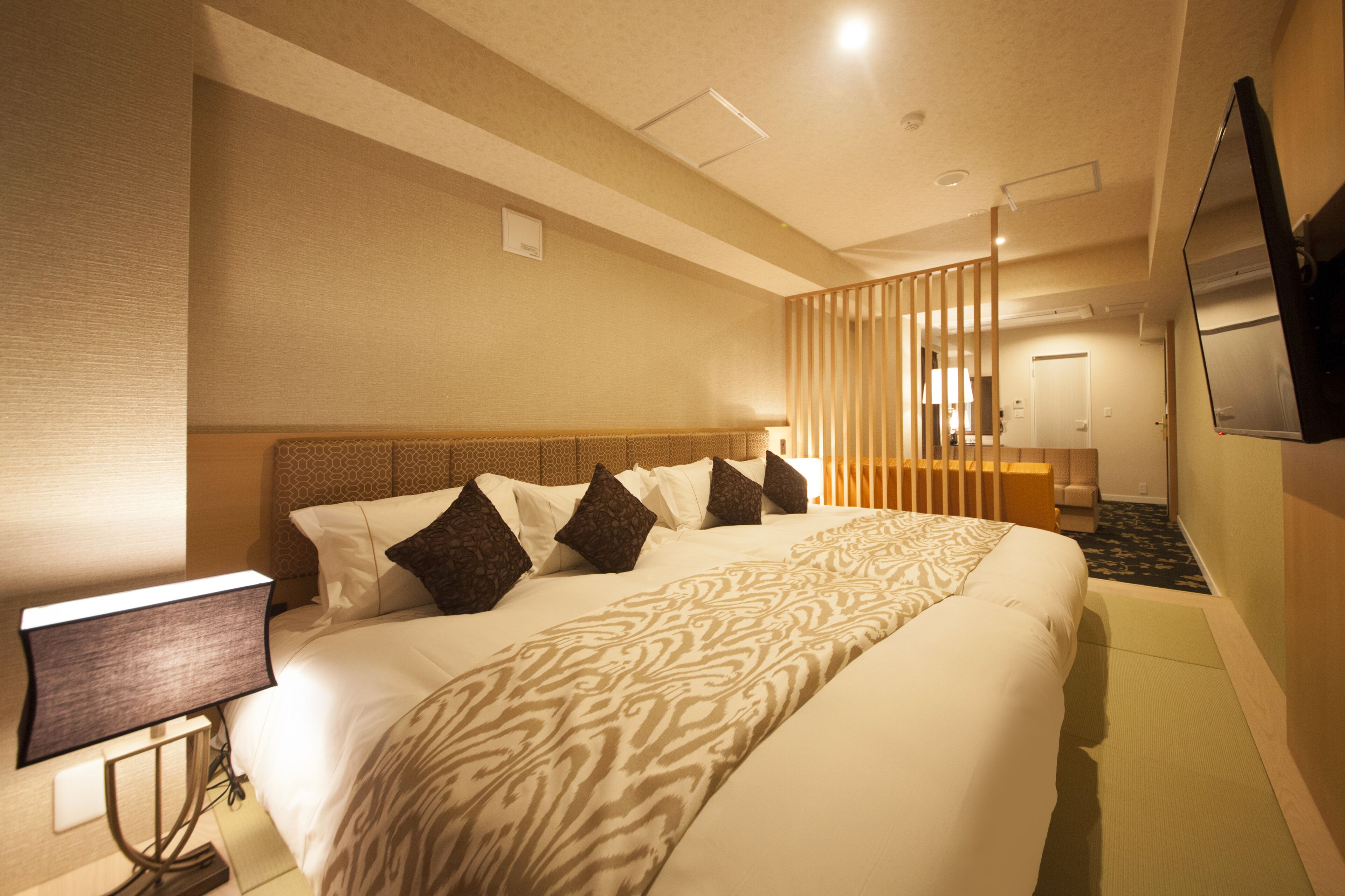 A hotel room with a large bed, a bedside table with a lamp, a TV mounted on the wall, and a wooden partition separating the room from the hallway.