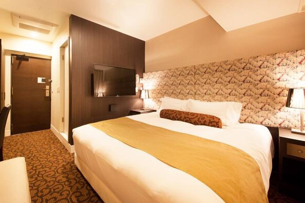 A hotel room with a bed, a desk, a TV, and a patterned headboard.