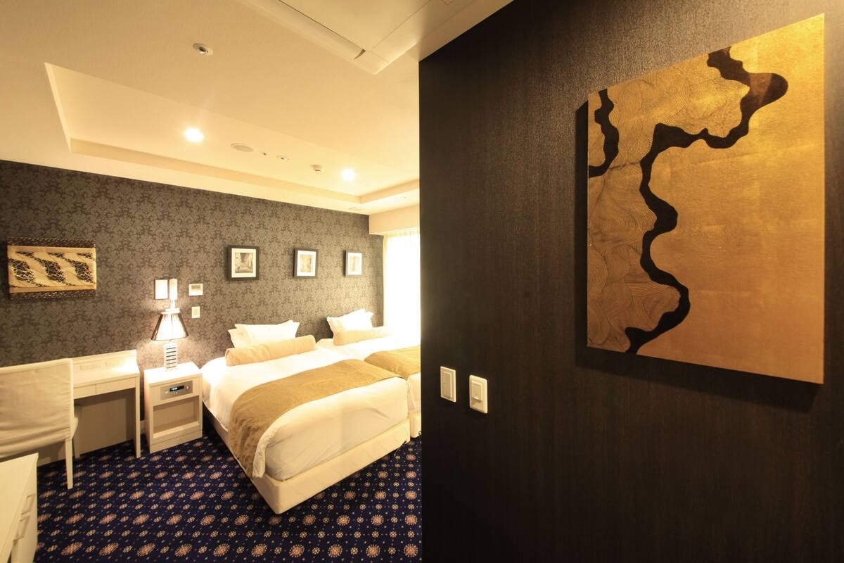 A hotel room with a bed, bedside tables, a desk, a lamp, framed pictures on the wall, and a large abstract painting.