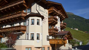Un hôtel de style alpin traditionnel, doté de balcons en bois et d’une enseigne indiquant les équipements disponibles, tels qu’un restaurant, une piscine d’été et un sauna.