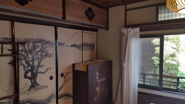 Una habitación tradicional japonesa con puertas correderas que exhiben una pintura de naturaleza, un armario de madera con relieves florales y una ventana con vistas a follaje.