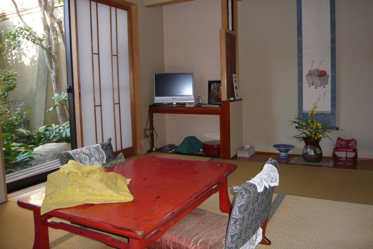 Una habitación japonesa tradicional con suelo de tatami, una mesa roja, un televisor sobre un soporte de madera y una puerta corredera shoji.