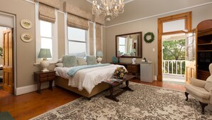 A bedroom with a large bed, bedside tables, a dresser, a chandelier, and a view through a door. - Royston Hall Guesthouse (Port Shepstone)