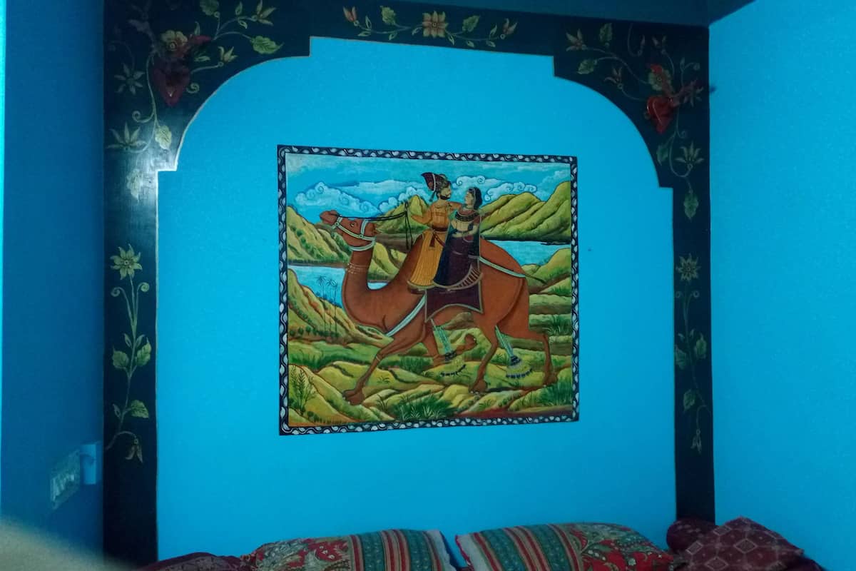 Une pièce décorée d’une fresque représentant un homme à cheval, avec un lit recouvert d’une literie à motifs et un mur bleu orné de motifs floraux.