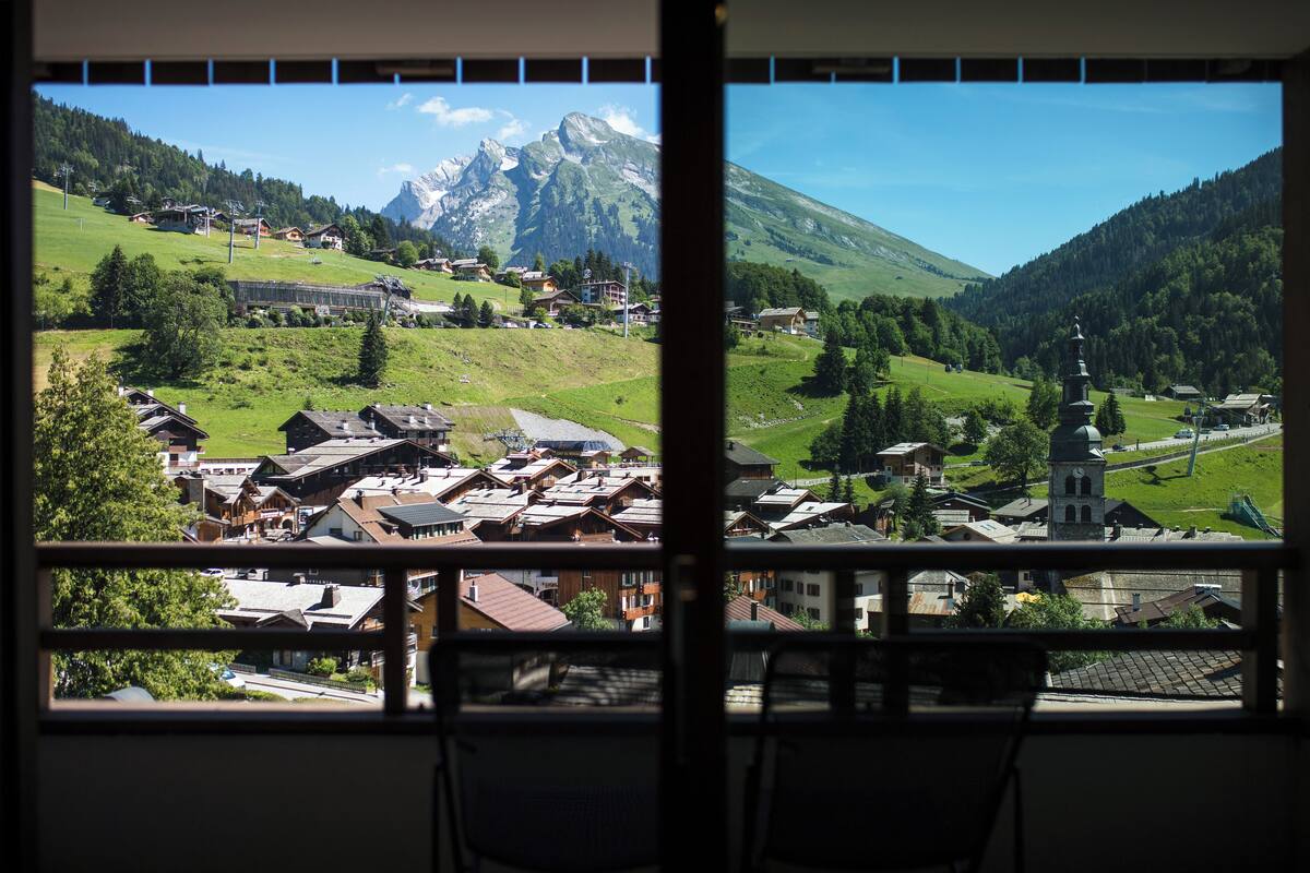 A view of a mountain village with traditional buildings and a church, seen through a window with a balcony.