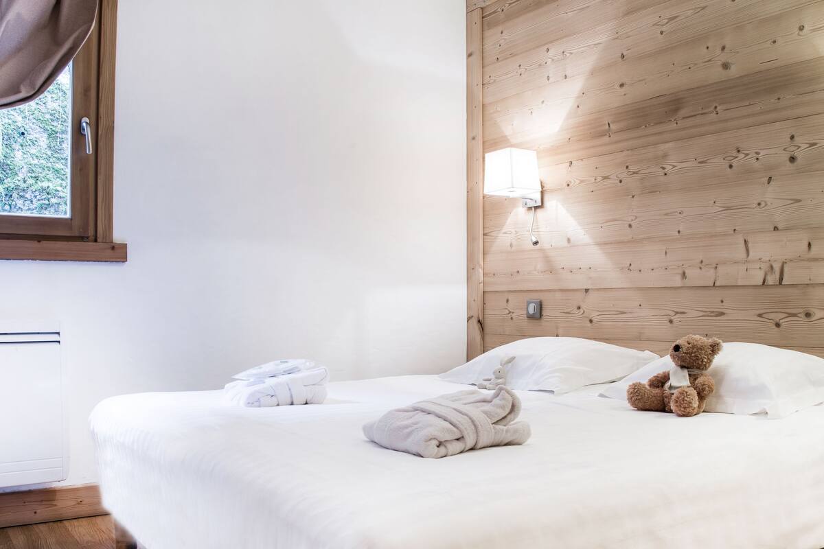 A neatly made bed with towels and a teddy bear, wooden panel walls, and a bedside table with a book.