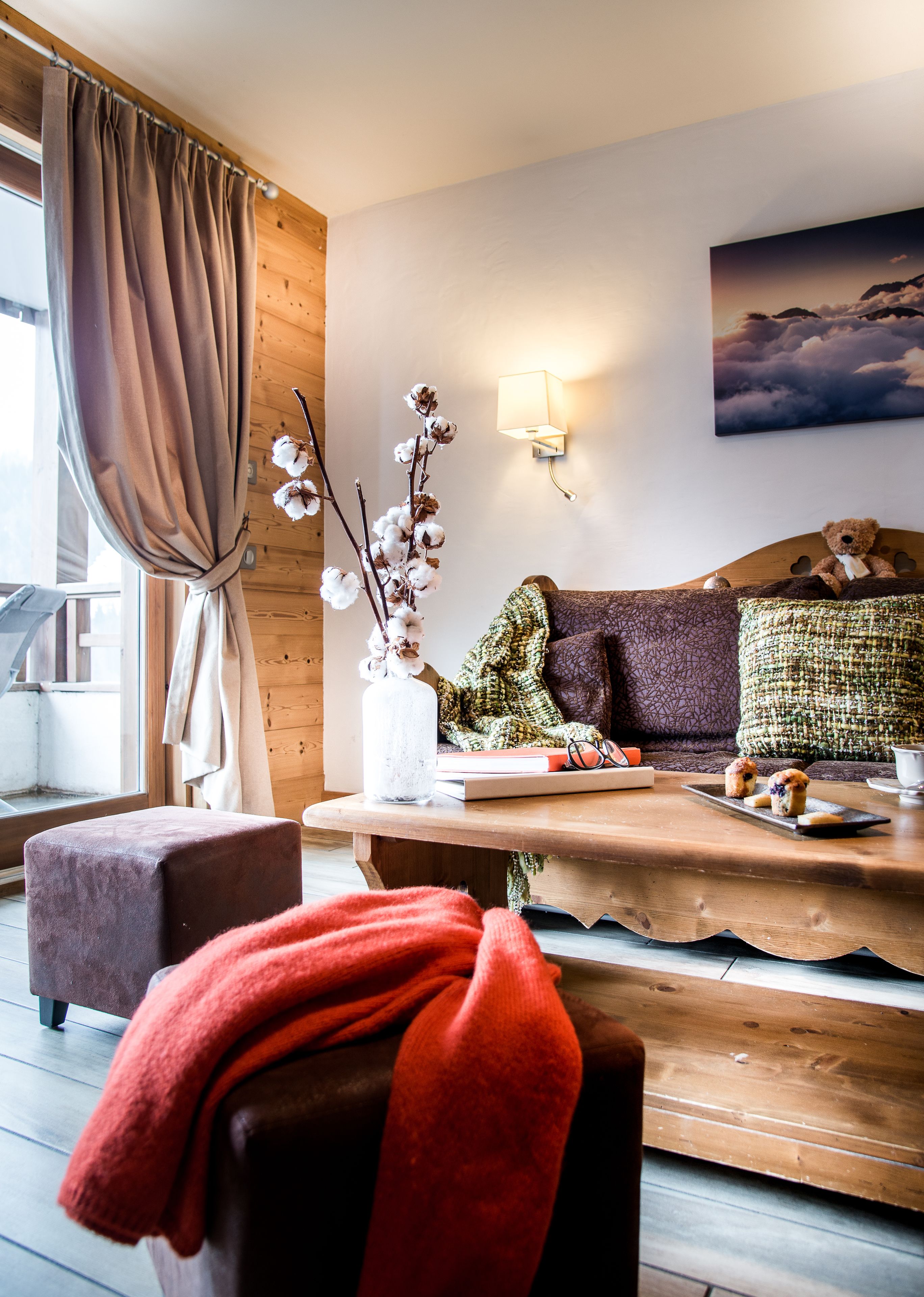 Un salon chaleureux avec un plancher en bois, un canapé agrémenté de coussins à motifs, une table avec des collations, une lampe et une fenêtre avec des rideaux.