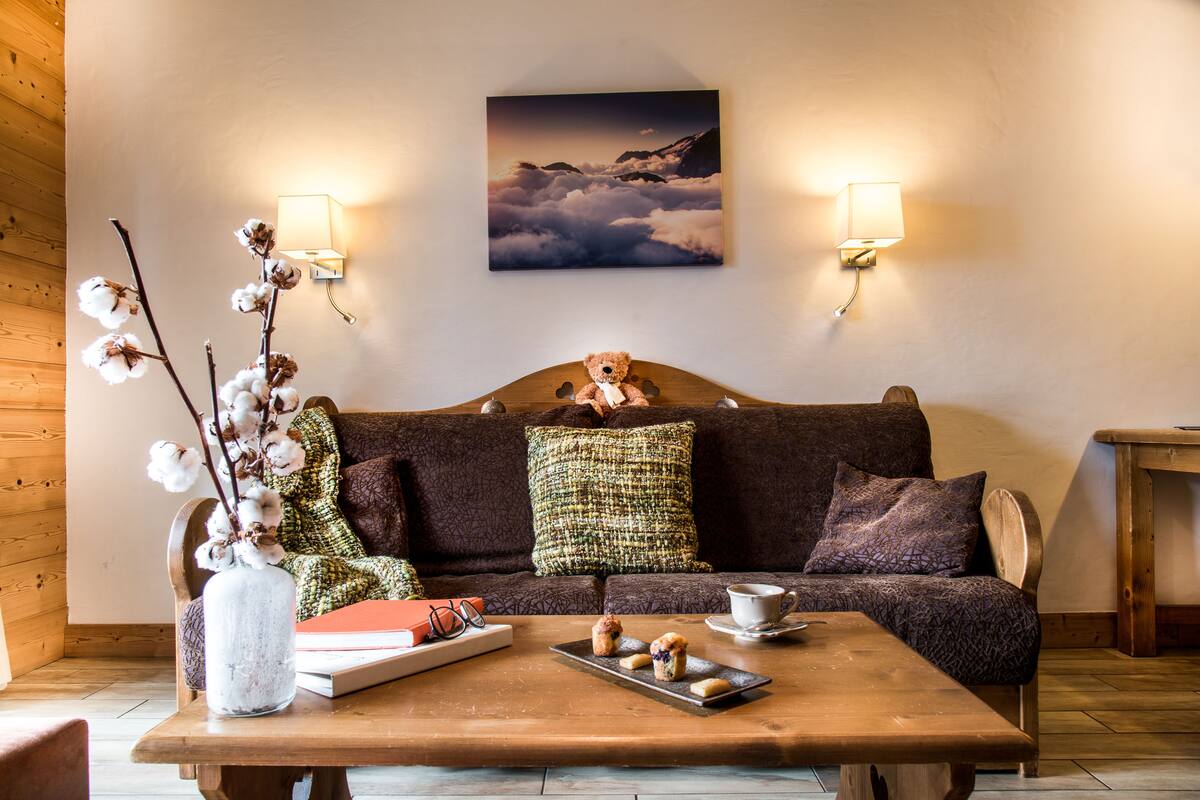 A cozy living room with a wooden floor, a brown sofa with patterned cushions, a vase with cotton branches, a tray with pastries, and a painting of clouds on the wall.