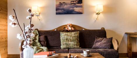 A cozy living room with a wooden floor, a brown sofa with patterned cushions, a vase with cotton branches, a tray with pastries, and a painting of clouds on the wall.