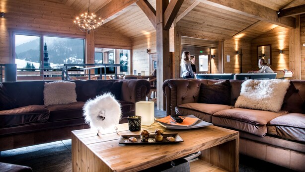 Interior acogedor de una cabaña con paredes y techo de madera, un ventanal con vistas a la montaña, un sofá de cuero marrón, una mesa de centro de madera con pasteles, y un perro blanco y esponjoso.