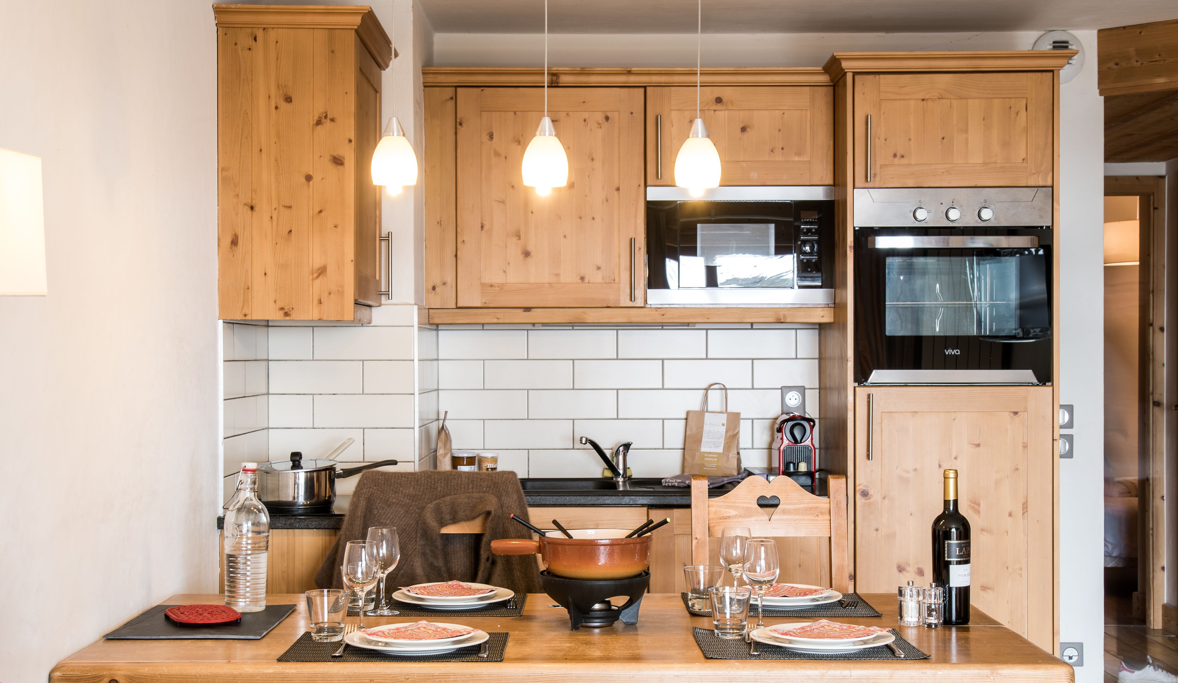A kitchen with wooden cabinets, a stove, and a dining table set for two with plates, glasses, and a bottle of wine.