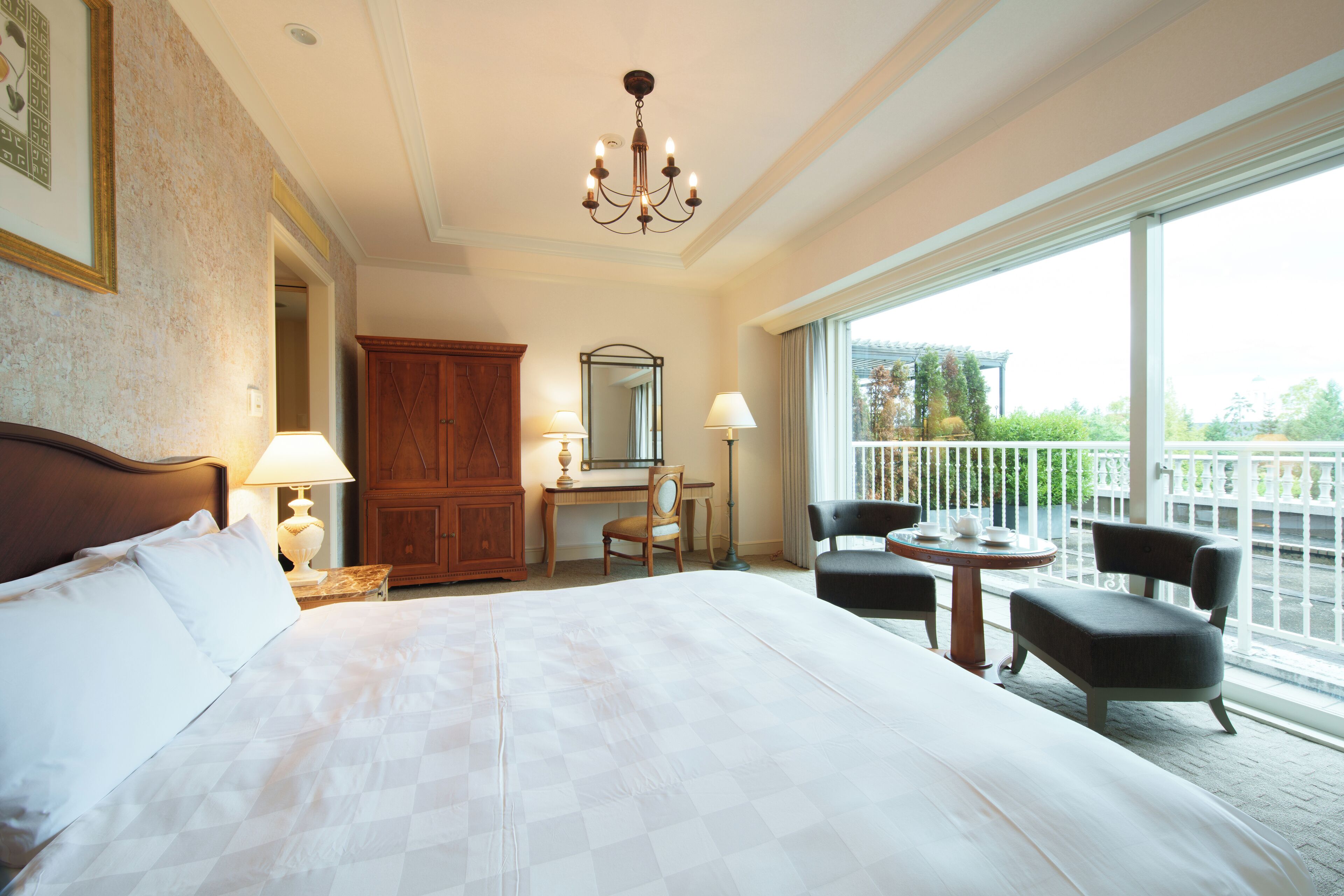 A hotel room with a large bed, a desk, a chair, a balcony with chairs, and a chandelier.