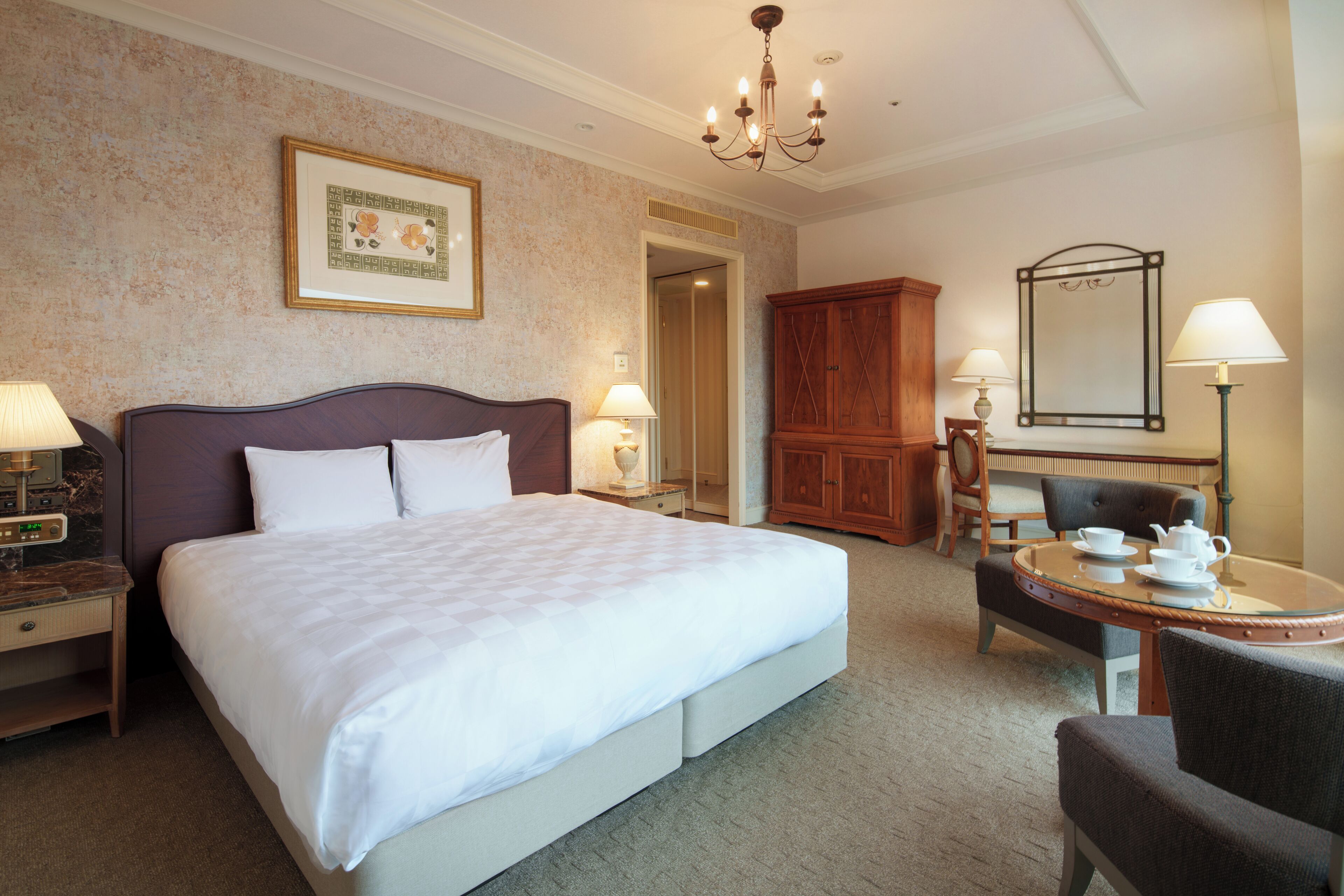 A hotel room with a large bed, a wooden cabinet, a round table with a chair, and a mirror.