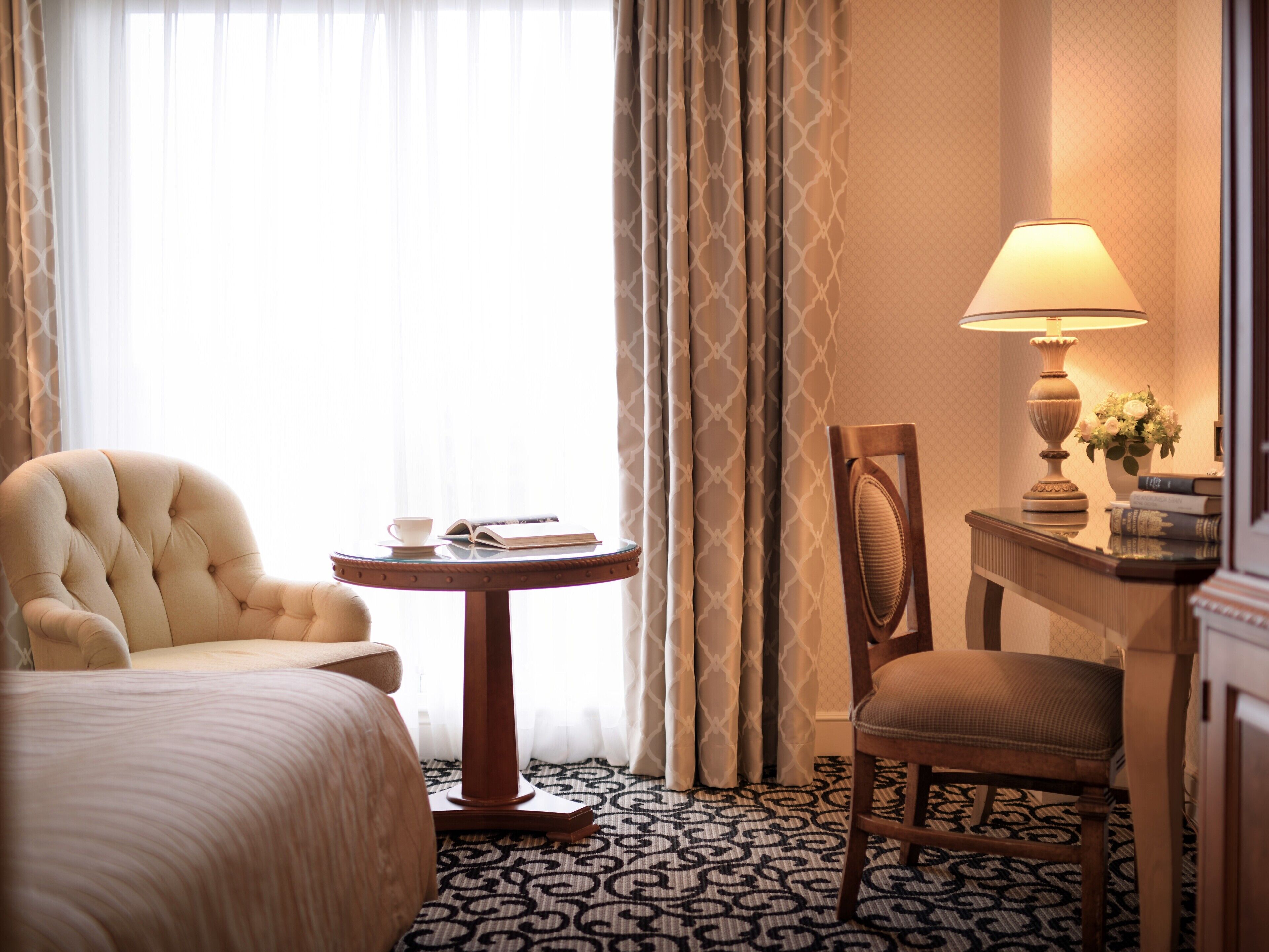A hotel room with a bed, a round table with a book and a cup, a chair, a lamp, and a small table with a vase of flowers.