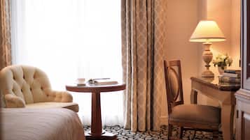 A hotel room with a bed, a round table with a book and a cup, a chair, a lamp, and a small table with a vase of flowers.