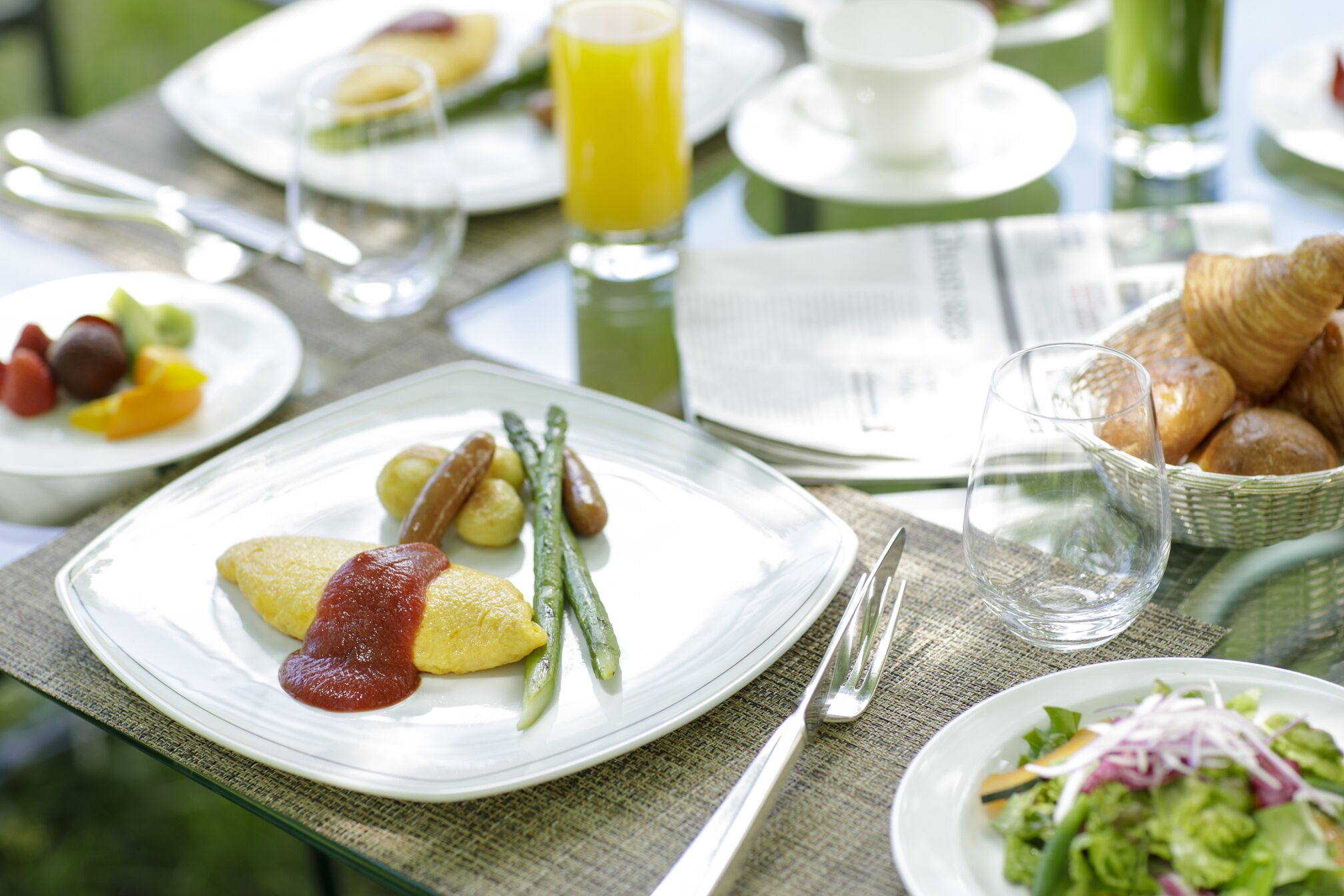 A breakfast setting with plates of food, a glass of orange juice, and a basket of croissants.
