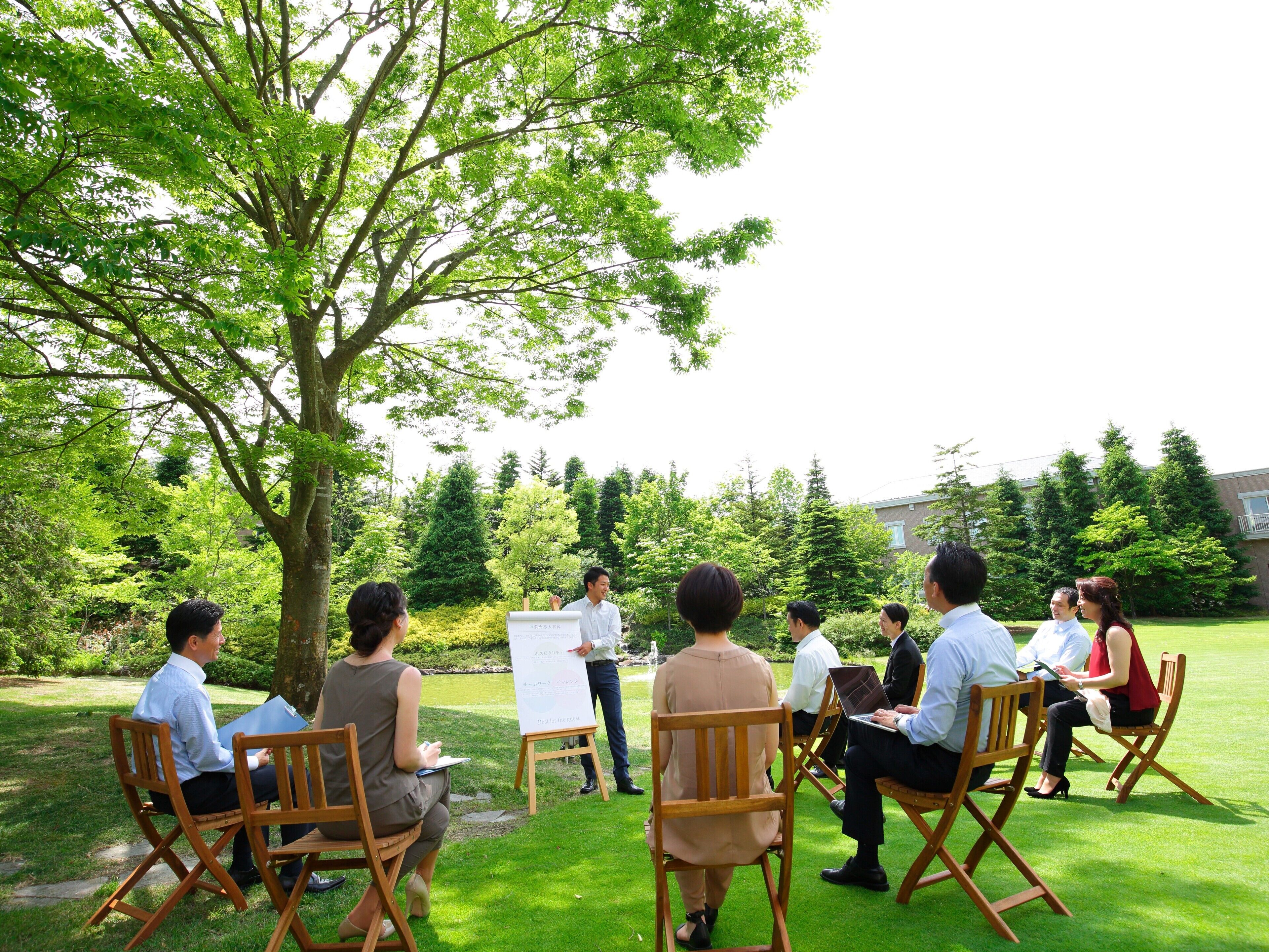 Un groupe de personnes est assis à l’extérieur, dans un espace verdoyant, avec des arbres et un bâtiment en arrière-plan.