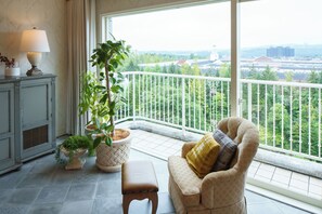 Un balcón con vista a un paisaje urbano, un sillón de mimbre con cojín, una mesita con lámpara y dos plantas en macetas.