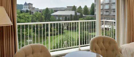 A balcony with two upholstered chairs and a small round table, overlooking a well-maintained garden and buildings in the background.