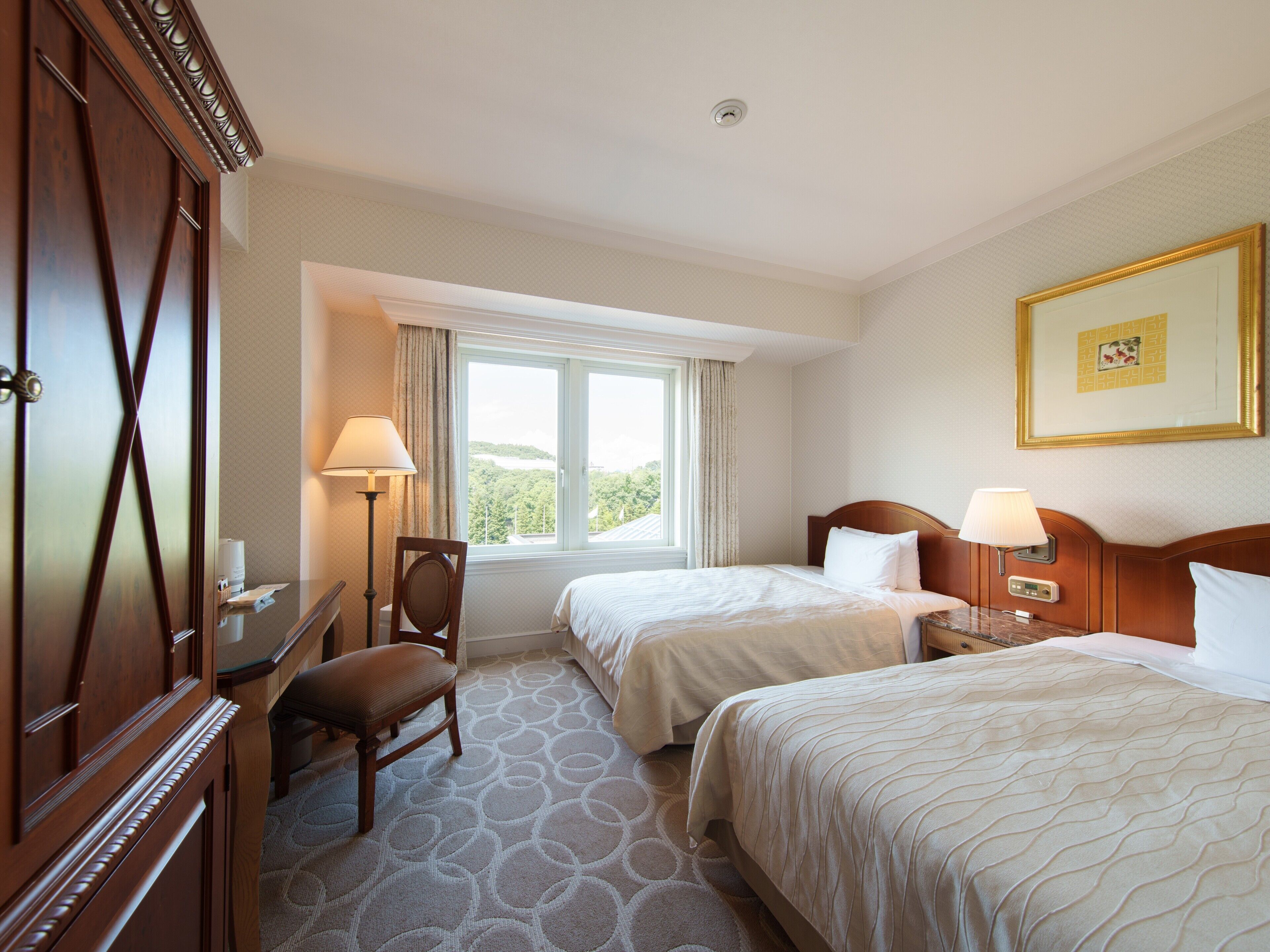 A hotel room with two beds, a desk, a chair, a lamp, and a window with a view.