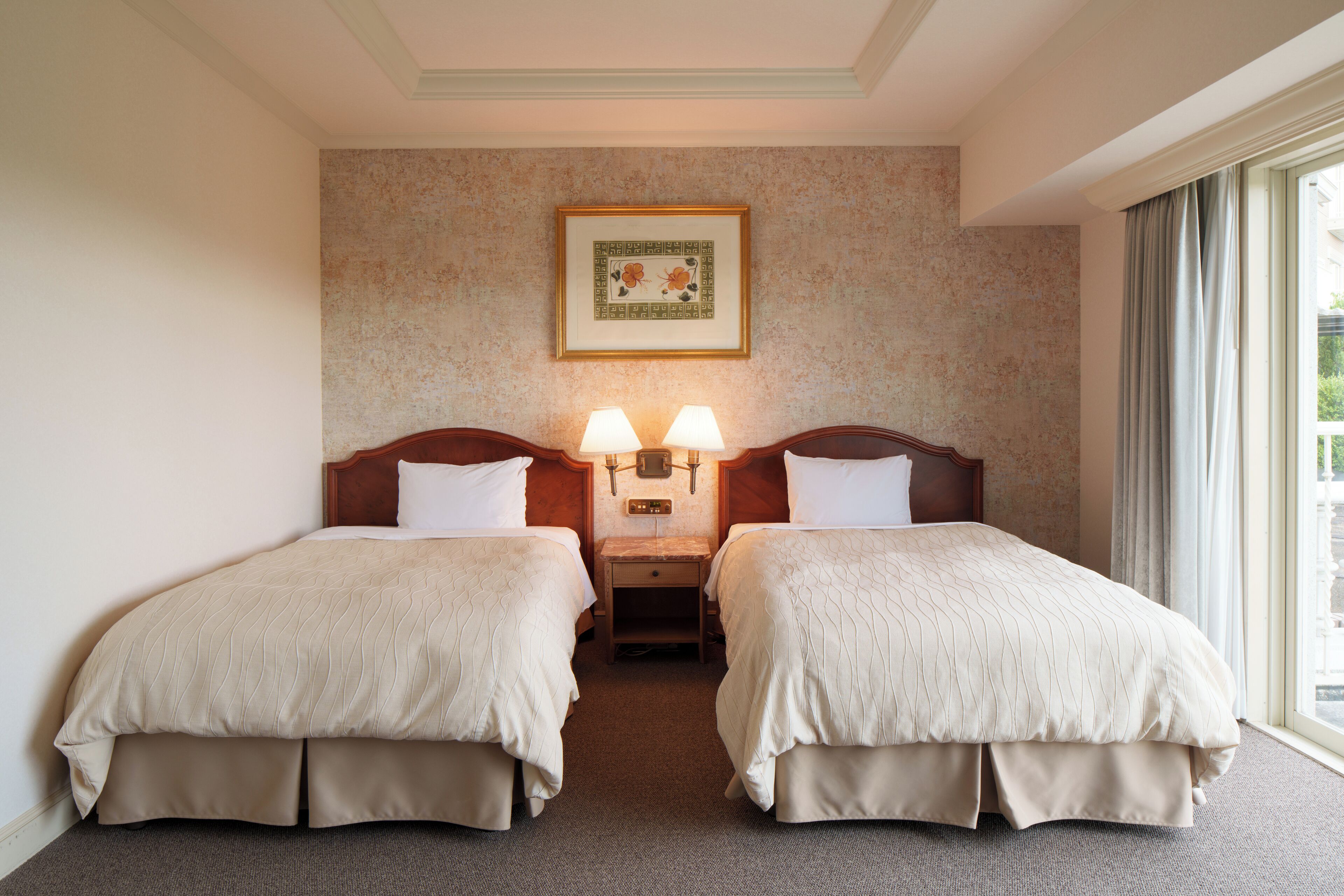 Habitación de hotel con dos camas, una mesita de noche con lámparas, un cuadro en la pared y una ventana con cortinas.