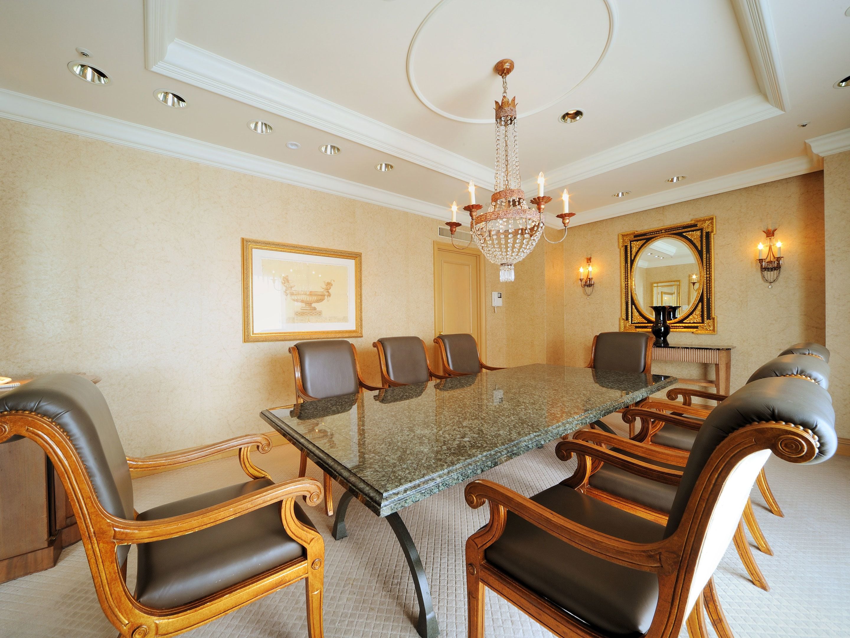 A formal meeting room with a glass-top table, leather chairs, a chandelier, and a large mirror on the wall.