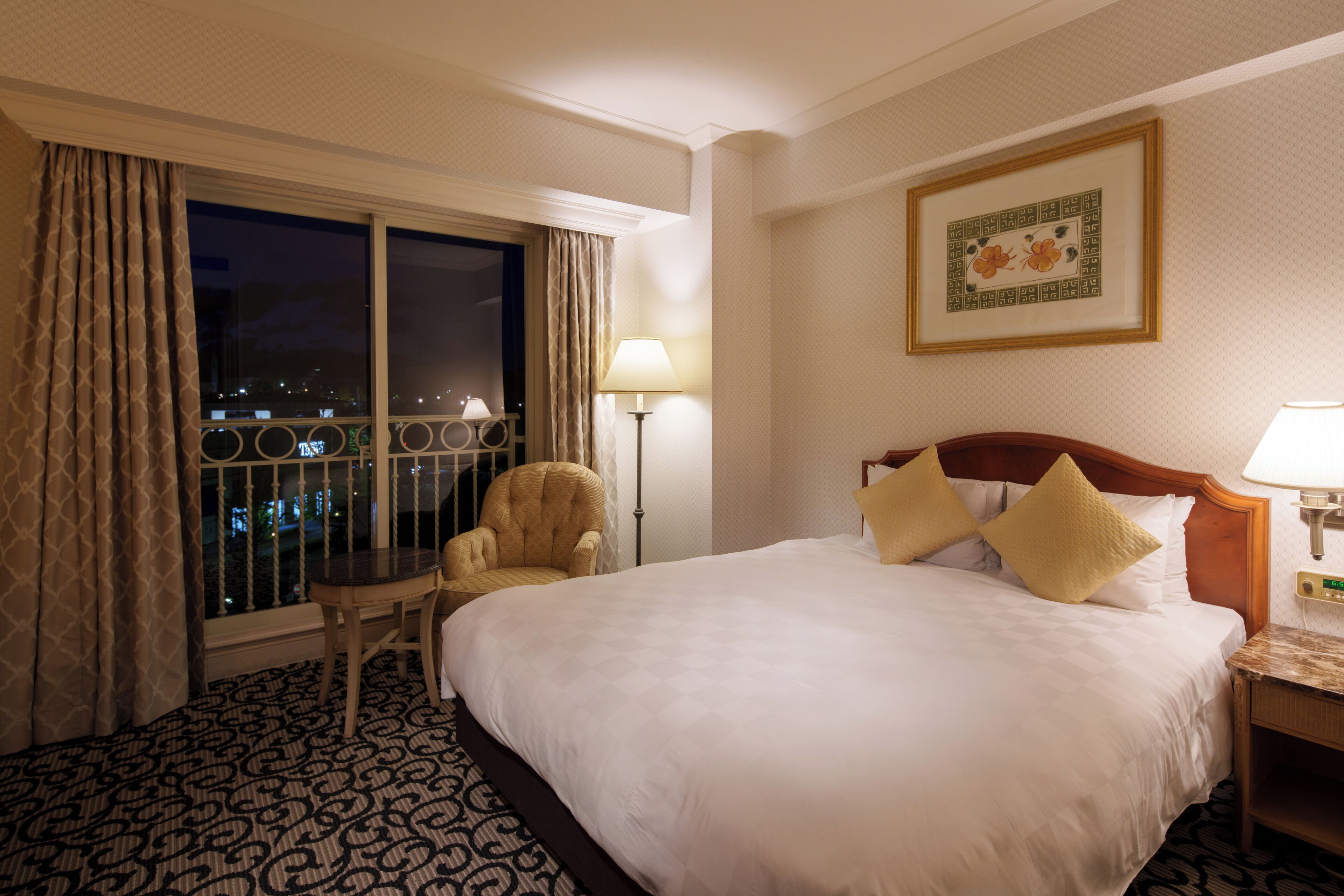 Une chambre d’hôtel avec un grand lit, une chaise, une table de chevet, une lampe, une fenêtre avec des rideaux et un tableau encadré au mur.