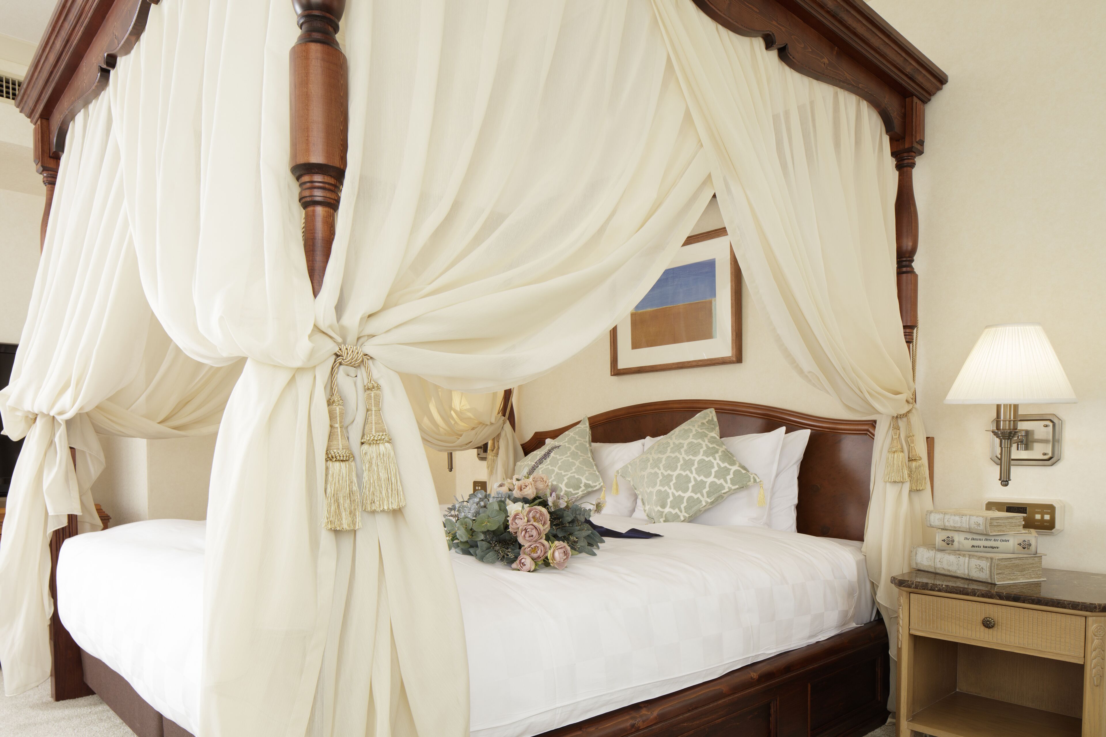 A four-poster canopy bed with a bouquet of flowers, a bedside lamp, and a framed picture on the wall.