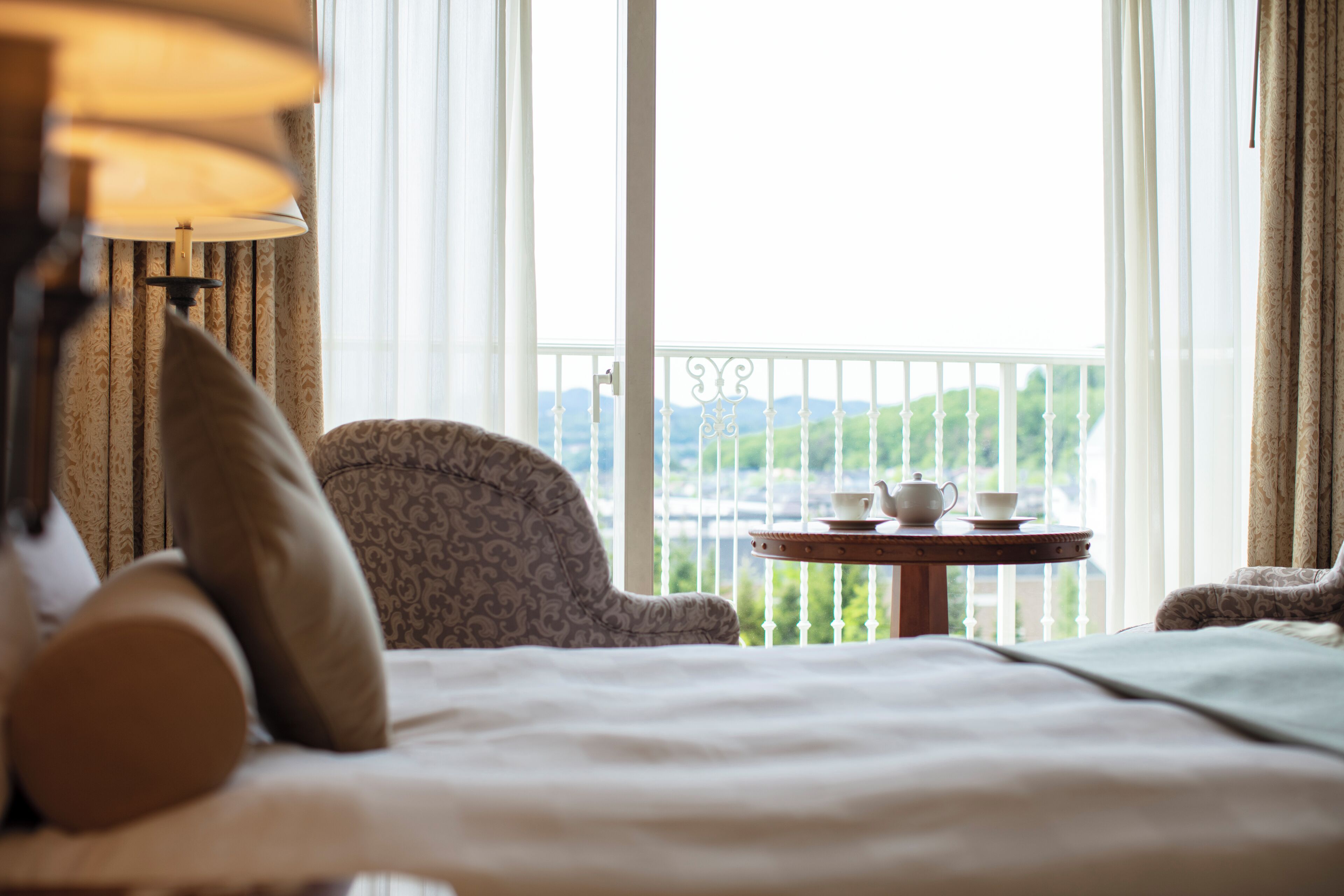 A hotel room with a bed, a chair, a table with a teapot and cup, and a view of greenery through the window.