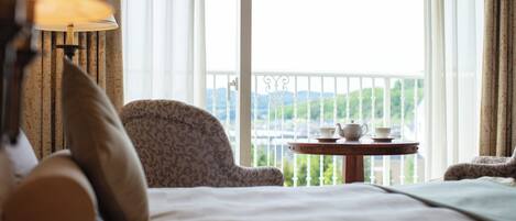 A hotel room with a bed, a chair, a table with a teapot and cup, and a view of greenery through the window.