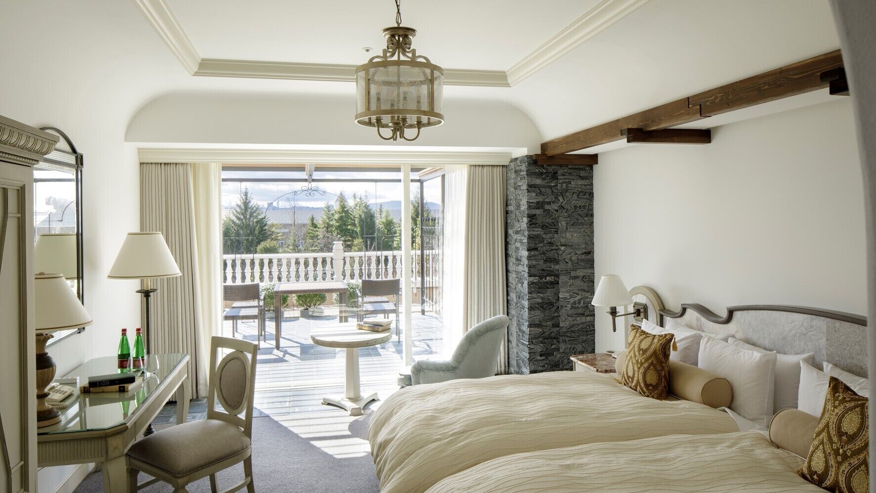 A hotel room with a bed, a desk, a chair, a small table, and a balcony with outdoor furniture.