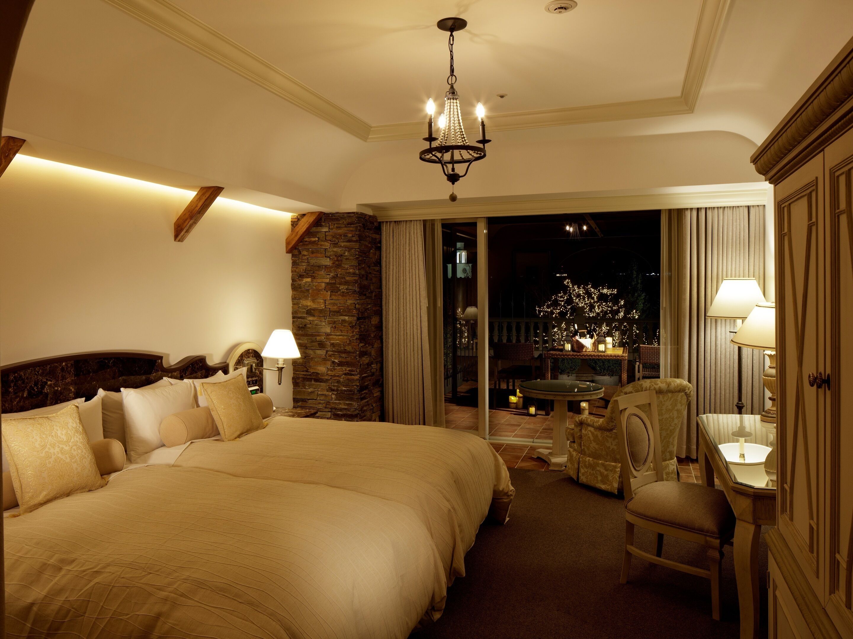 Amplia habitación de hotel con una cama grande, un muro decorativo de piedra, un candelabro y vistas del exterior por la noche.