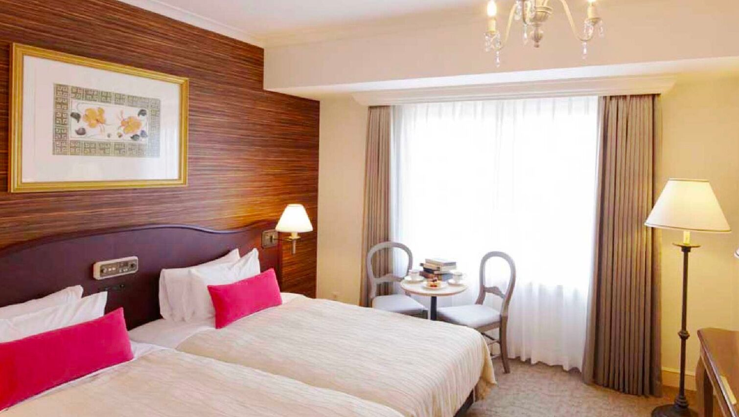 A hotel room with two beds, a small table with chairs, a chandelier, and a framed picture on the wall.