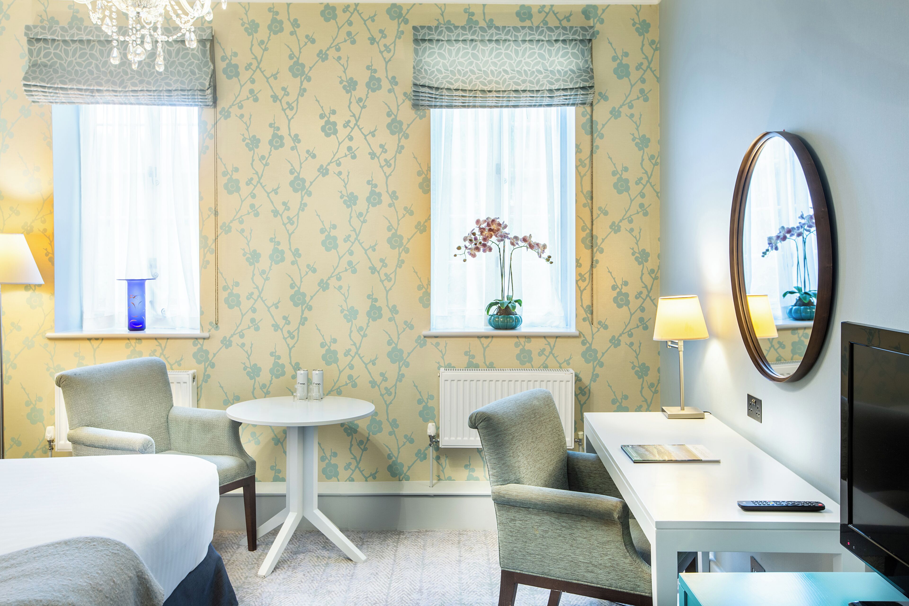 A hotel room with a bed, two armchairs, a small table, a TV, and a window with a plant.
