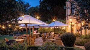 Un patio al aire libre con mobiliario de mimbre, sombrillas blancas y luces colgantes.