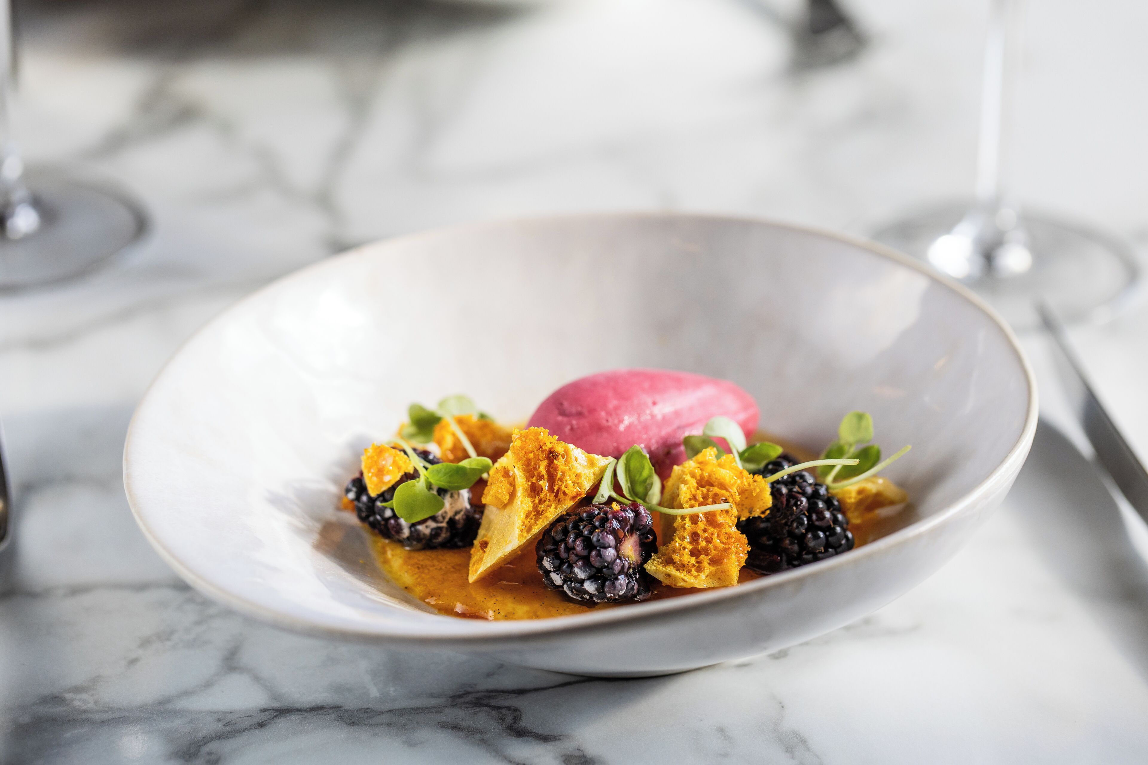 A dessert plate with a scoop of ice cream, blackberries, and honeycomb pieces on a marble surface.