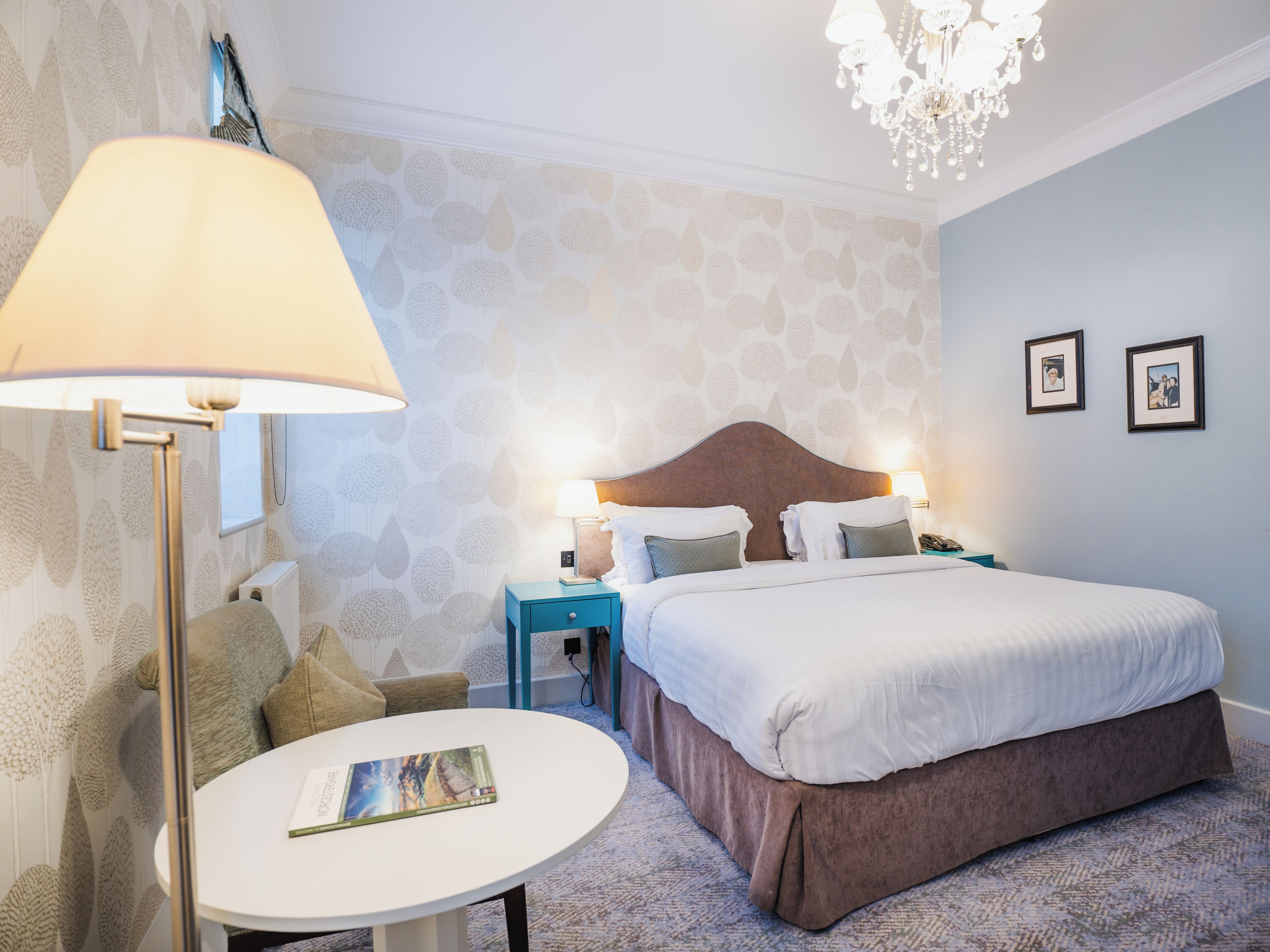 Hotellihuone, jossa on sänky, yöpöydät, lamppu, pyöreä pöytä ja kattokruunu.
