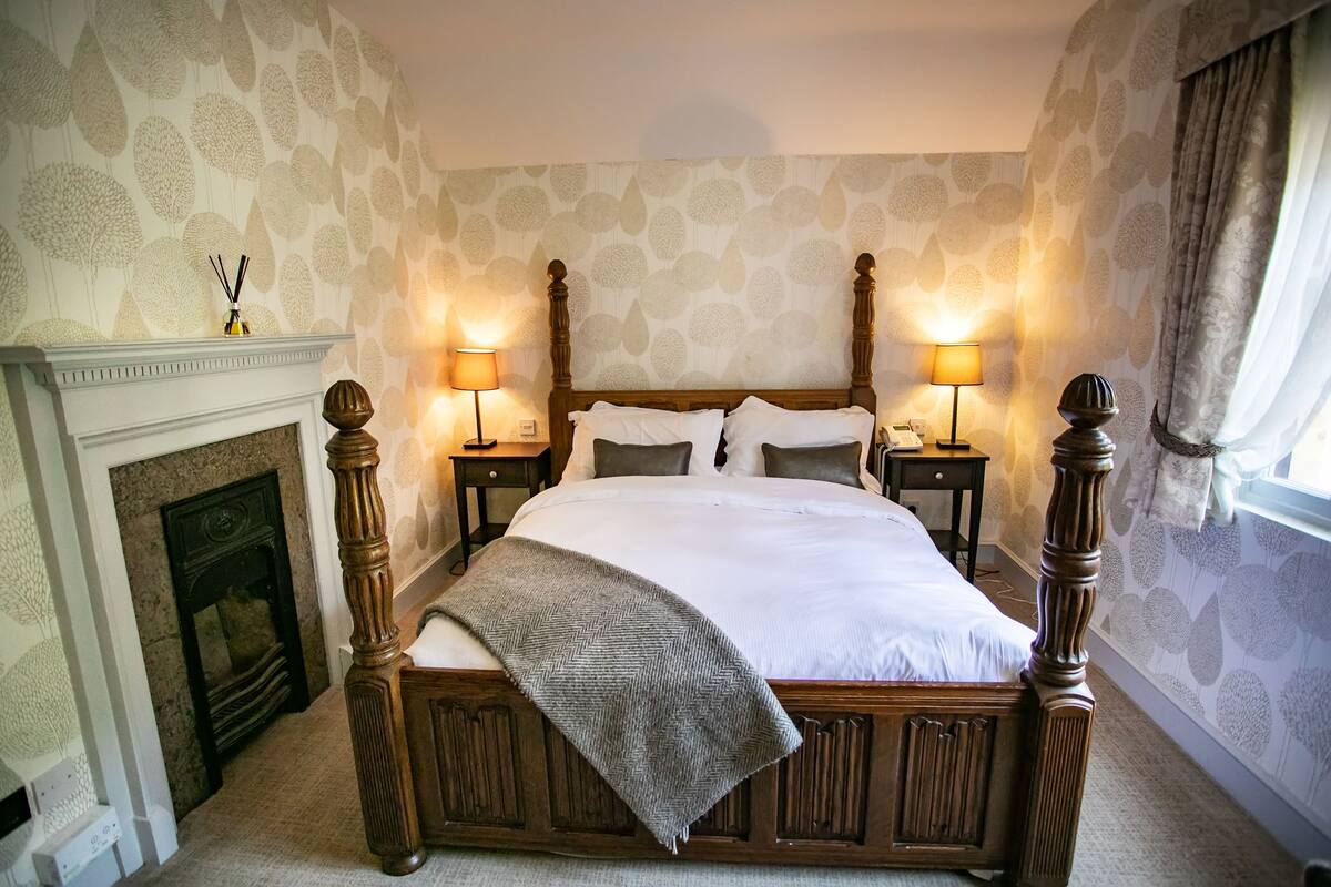 A bedroom with a large bed, two bedside tables with lamps, a fireplace, and patterned wallpaper.