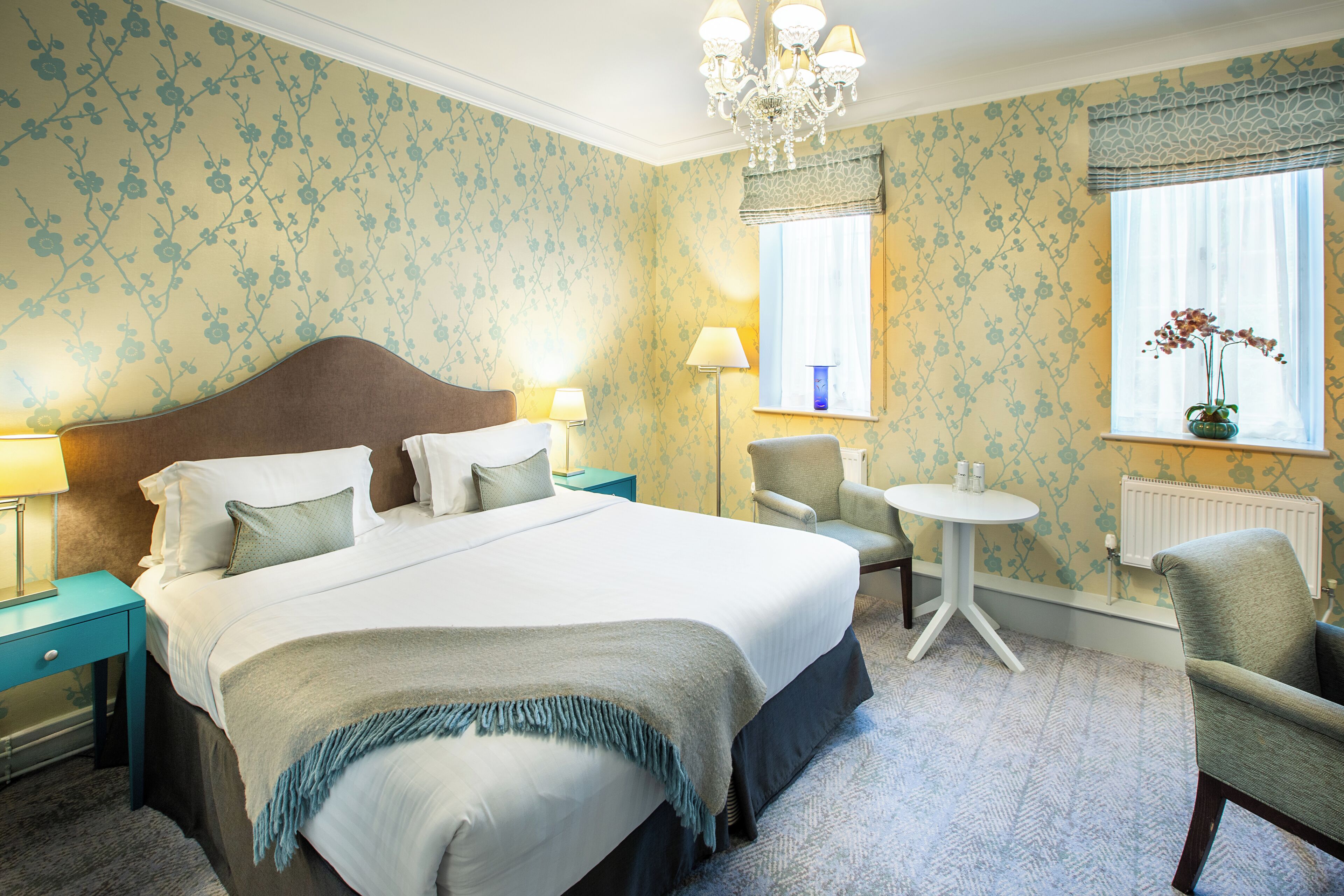 A hotel room with a large bed, two bedside tables, a chair, a small table, and a window with curtains.