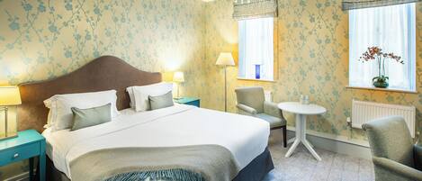 A hotel room with a large bed, two bedside tables, a chair, a small table, and a window with curtains.