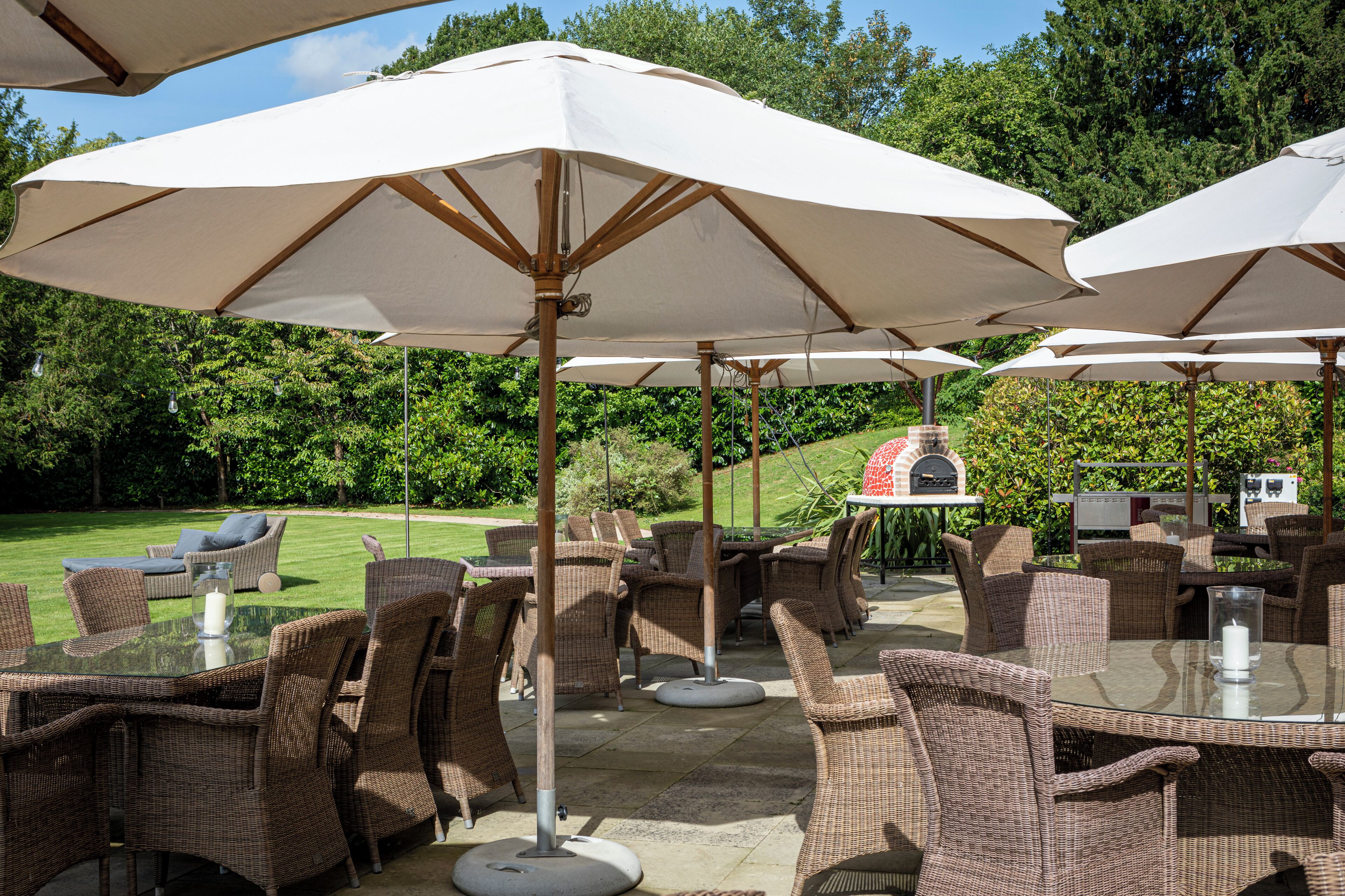 Outdoor wicker furniture with white umbrellas, a glass table, and a barbecue grill in the background.