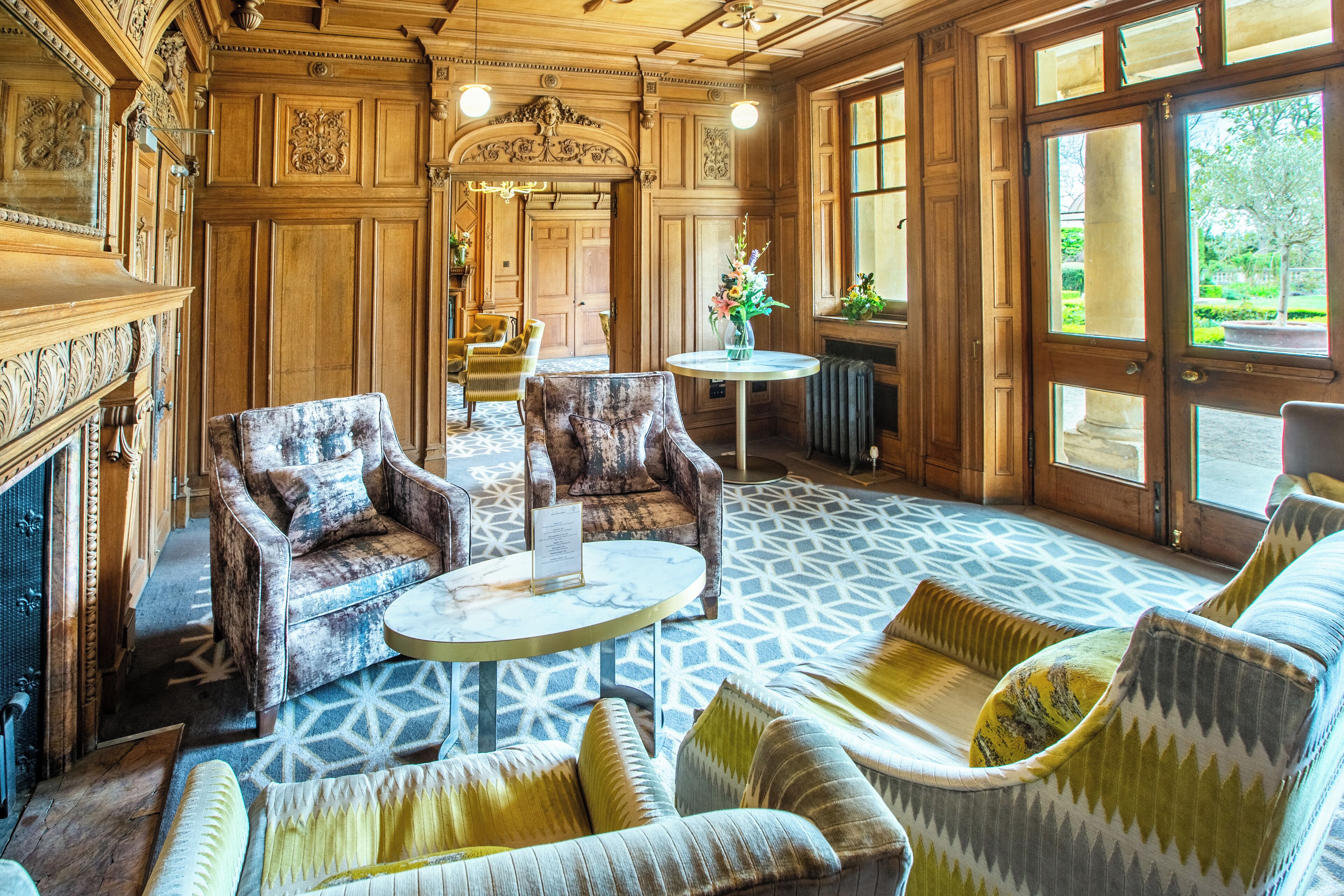 A spacious room with a patterned floor, wooden paneling, and a fireplace. There are several upholstered chairs and a round table with a vase of flowers.