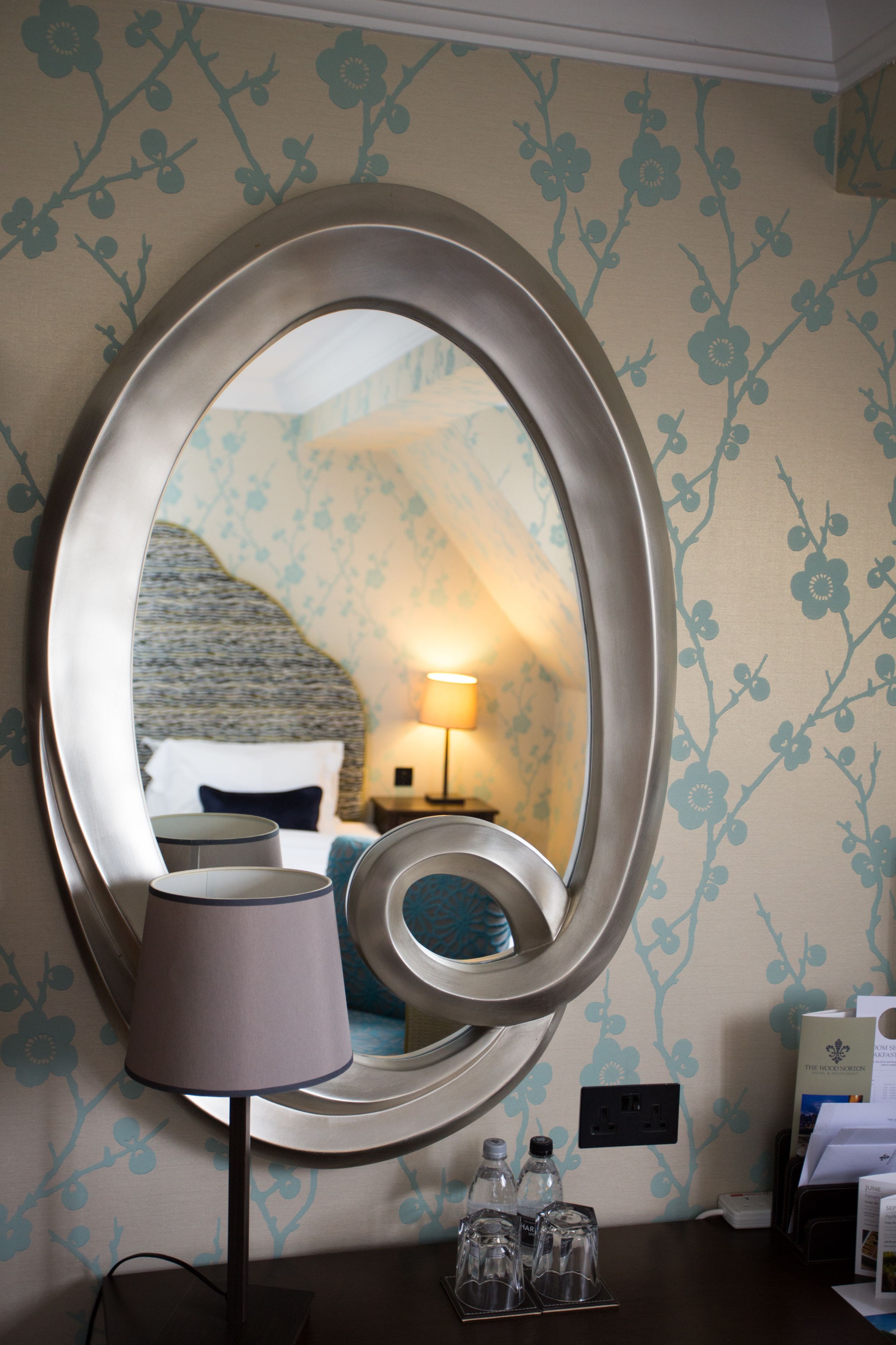 A mirror with an oval frame, a bedside lamp, and a small table with glasses and bottles.