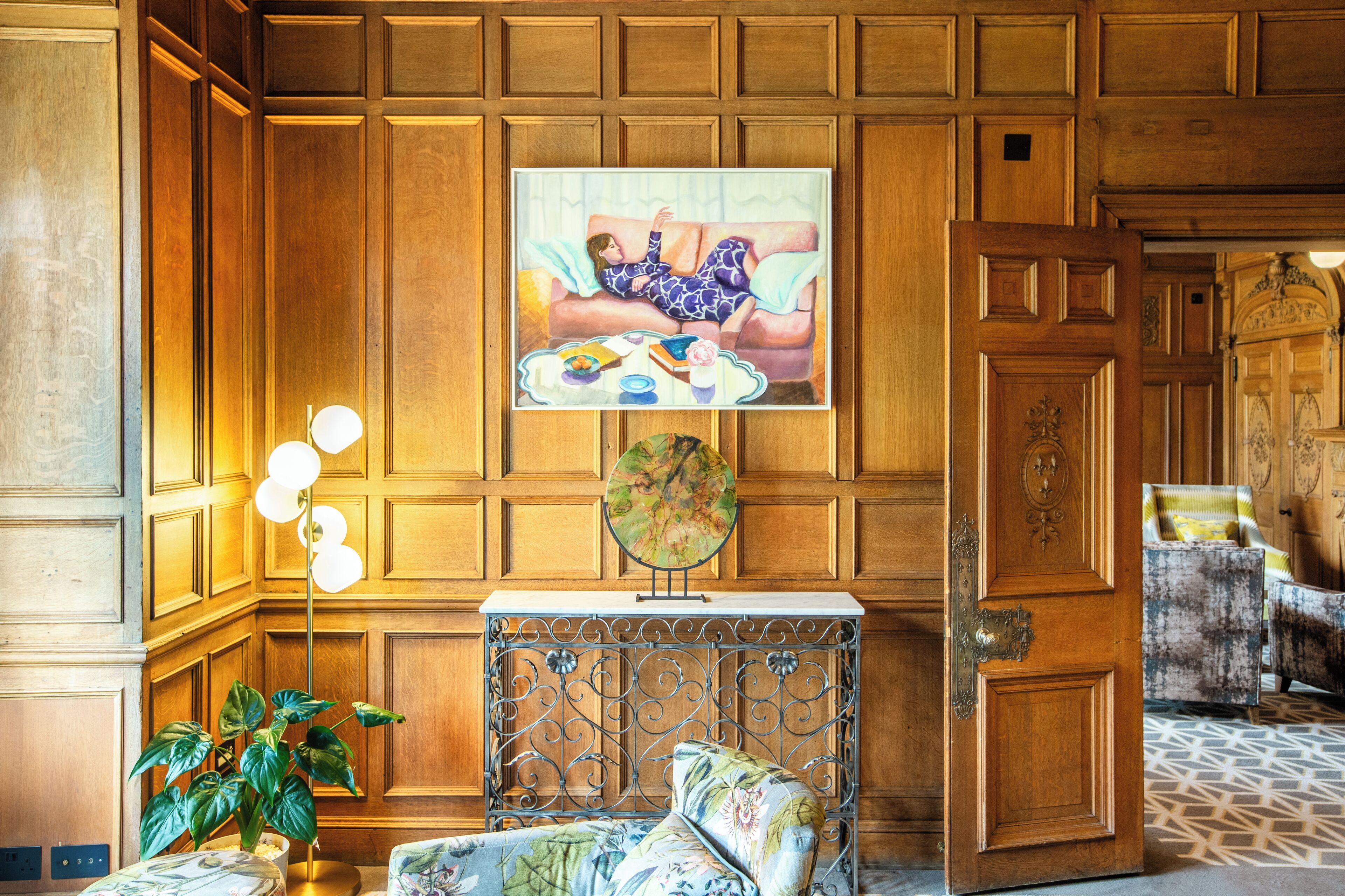 Een kamer met een houten lambrisering, een schilderij van een vrouw op een bank, een ronde tafel met een bloemenkleed en een potplant.