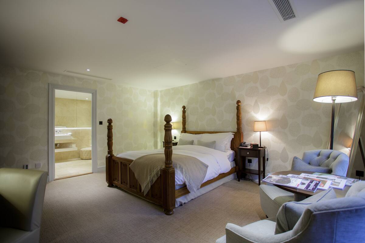 A hotel room with a four-poster bed, a bedside table with a lamp, a chair, and a small table with magazines.