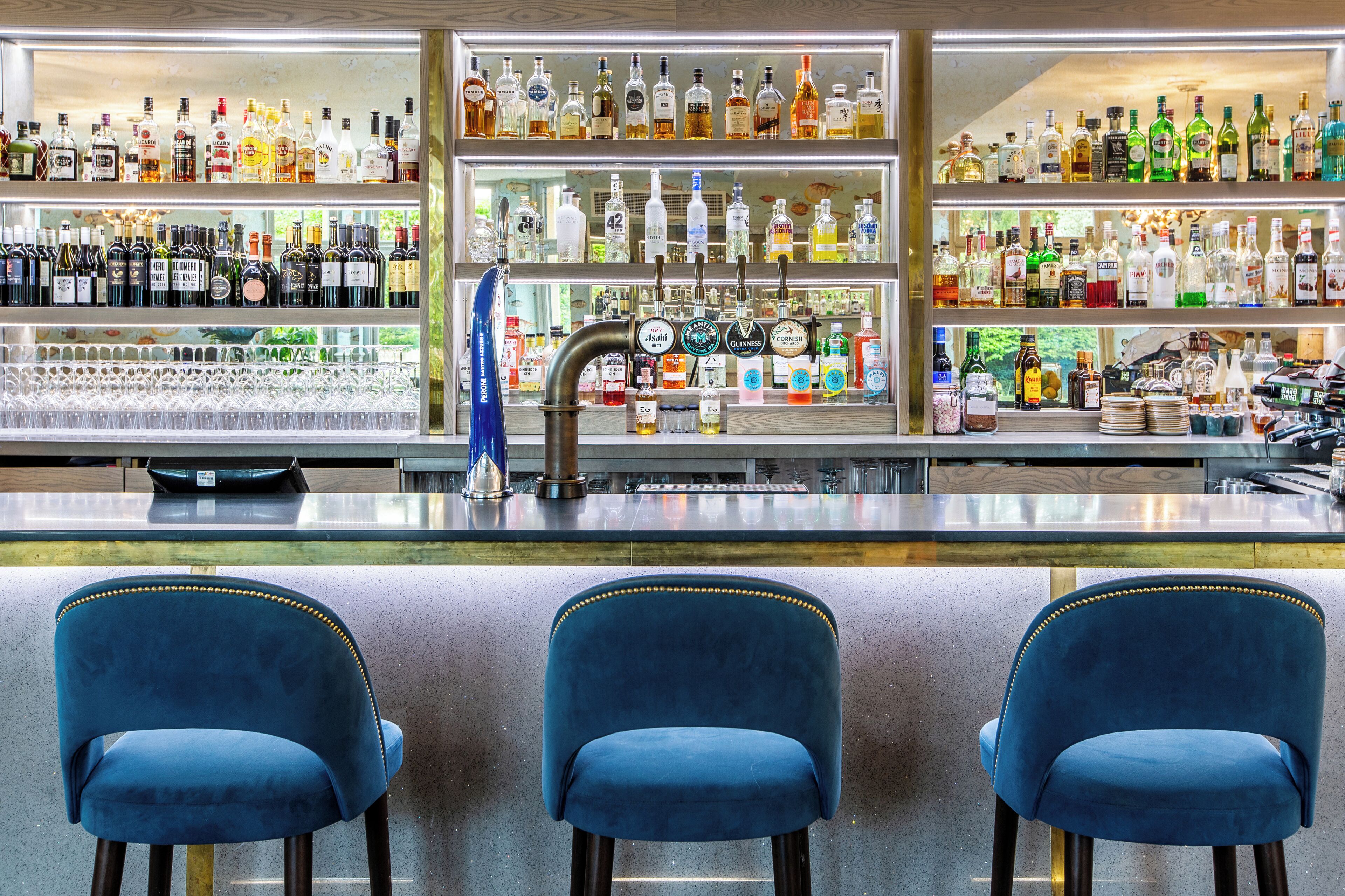 A bar area with multiple shelves of liquor bottles, a tap bar, and three blue chairs.